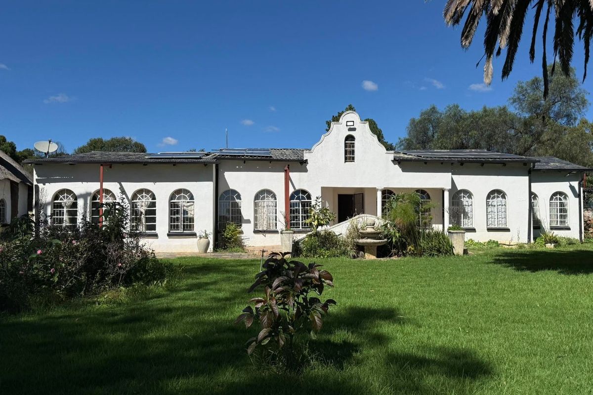 A well-maintained farm-style residence featuring a white facade, arched windows, solar panels, and a central stone fountain set within a spacious green garden.