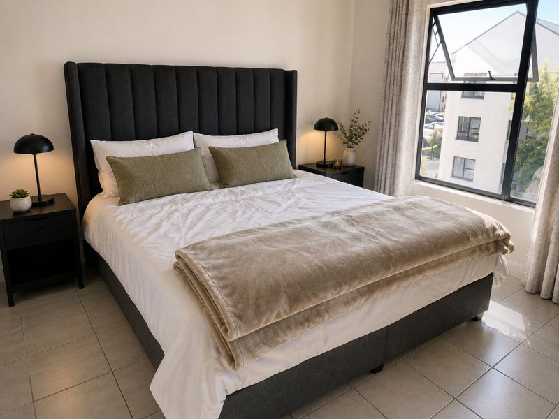 A well-lit bedroom featuring neutral tiled flooring and a large window with curtain treatments.