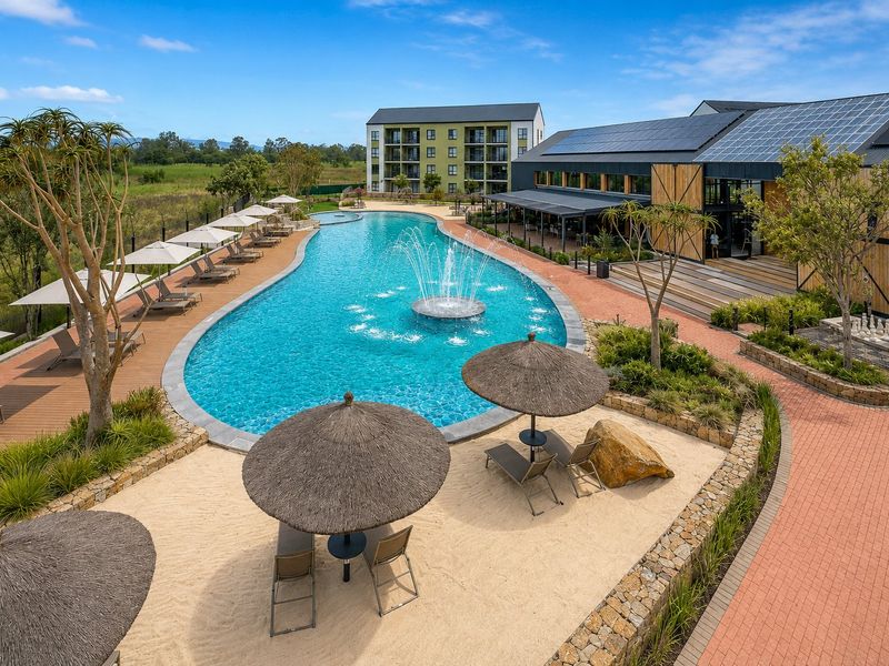 A well-maintained communal pool area featuring a central water fountain, paved walkways, landscaped surroundings, and solar panels on the adjacent building roof.