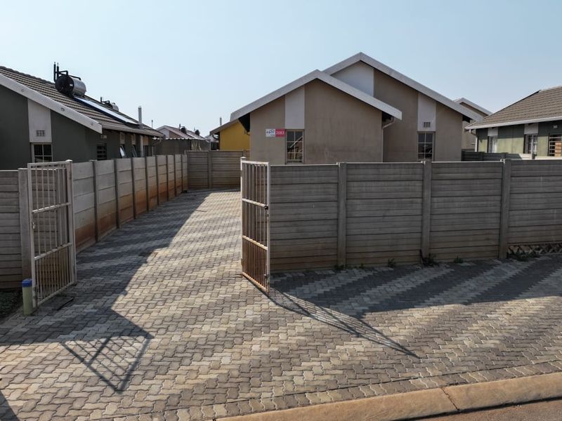 A front view of a residential property featuring a paved driveway, enclosed by a precast concrete boundary wall with a metal swing gate.