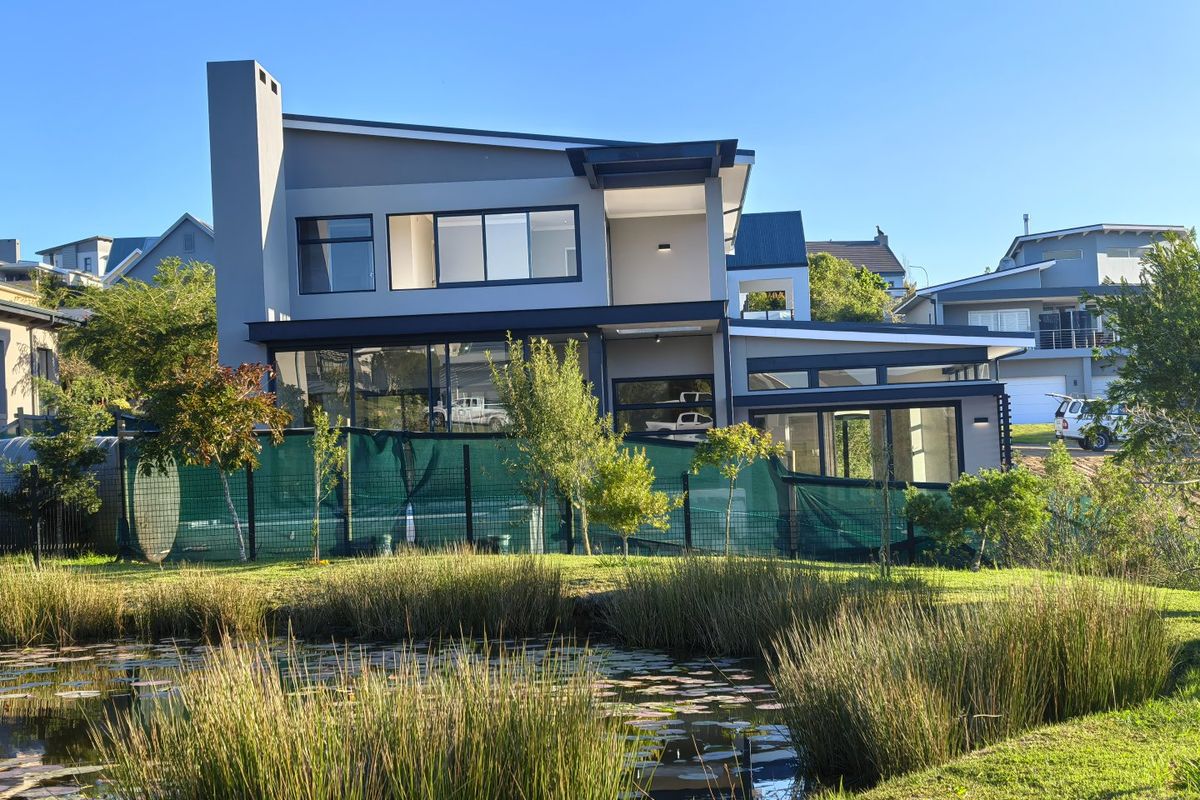 A modern double-story residence featuring large glass windows, a prominent chimney, and a covered patio overlooking a natural pond and landscaped garden.