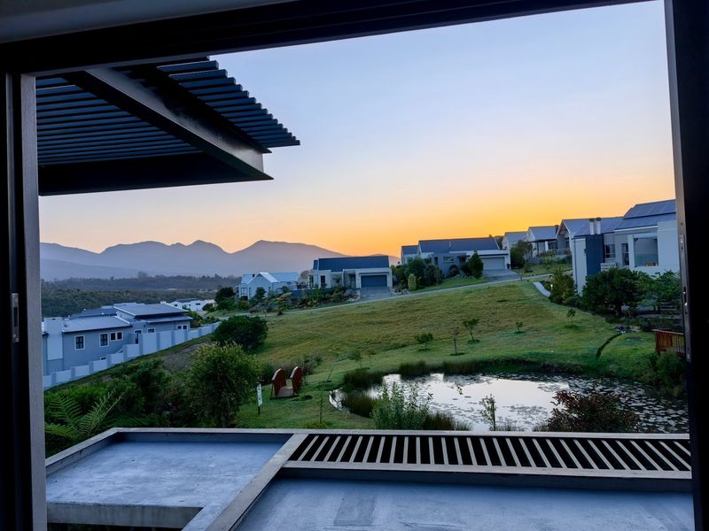 Scenic sunset view overlooking a landscaped estate with a pond, rolling lawns, and distant mountain ranges, framed from a modern balcony.