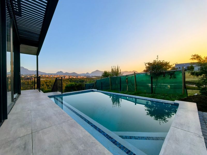 A modern swimming pool area with a tiled patio, metal pergola, and scenic mountain views at sunset.