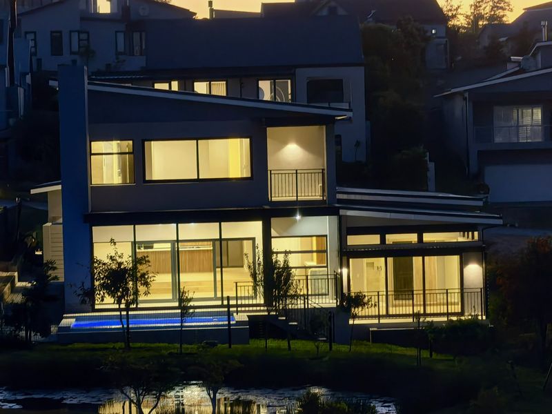 A modern multi-level house exterior at dusk, featuring large glass sliding doors, a swimming pool, and illuminated balconies.