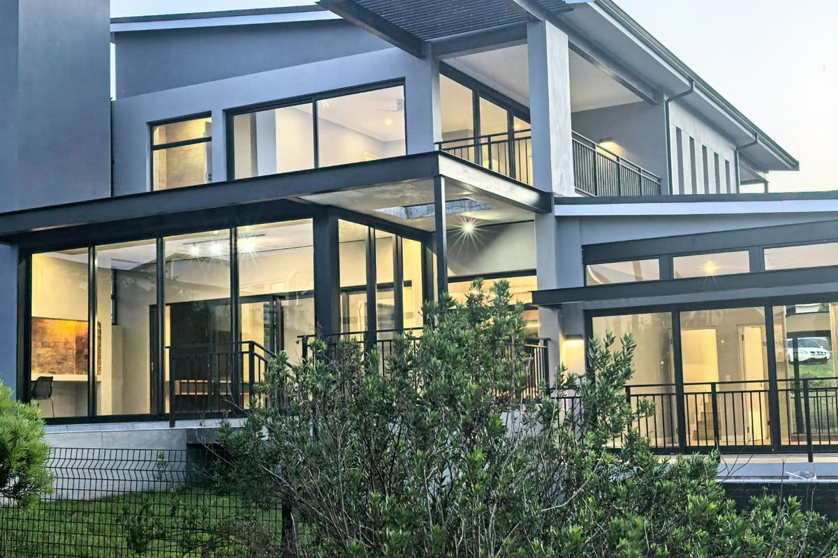 A modern multi-level rear facade featuring extensive glass sliding doors, a balcony with metal railings, and a contemporary steel pergola structure.