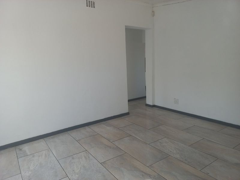 An empty living area featuring neutral tiled flooring, white walls, and dark grey skirting boards, providing a clean and versatile space.