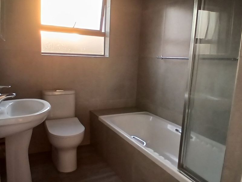 A clean, functional bathroom featuring a pedestal basin, toilet, and a built-in bathtub with a glass shower screen and neutral wall tiling.