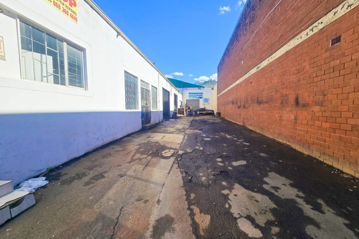 Rear industrial access lane featuring asphalt paving between a white painted building and a red brick boundary wall.