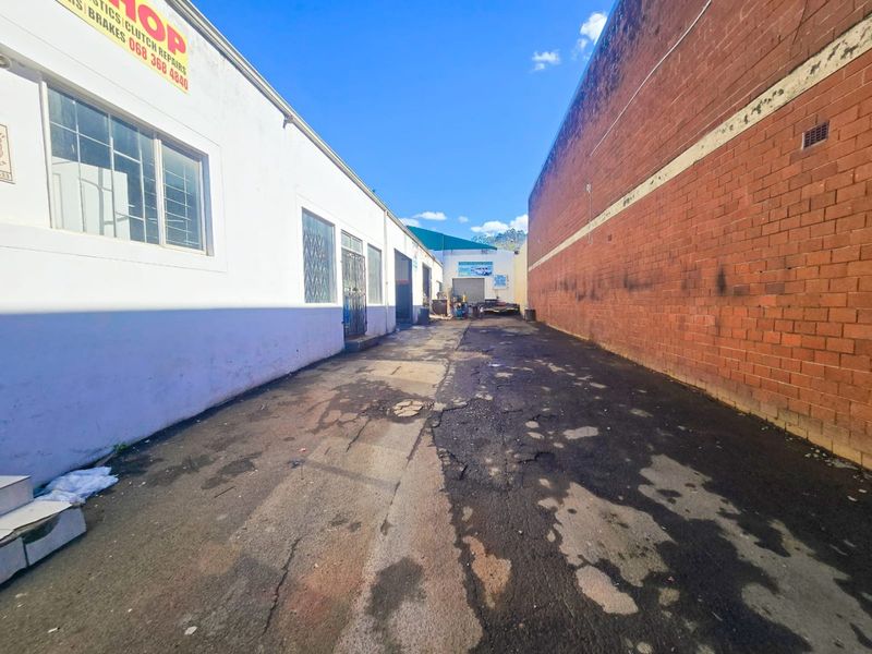 Rear industrial access lane featuring asphalt paving between a white painted building and a red brick boundary wall.