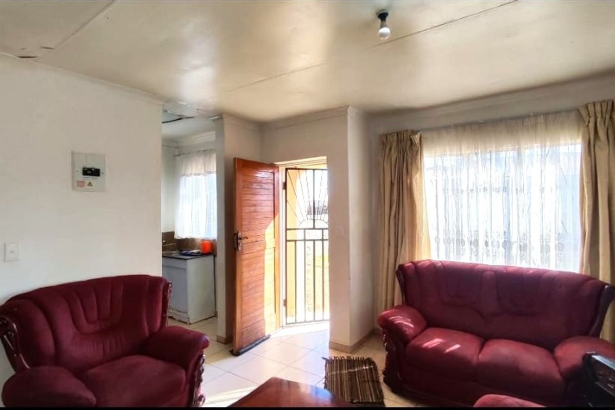 A living room area featuring tiled flooring, a security gate, and access to an adjacent kitchen space, showing signs of wear on the ceiling.