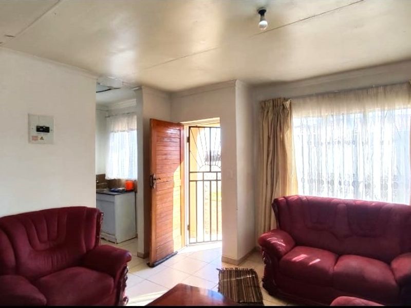A living room area featuring tiled flooring, a security gate, and access to an adjacent kitchen space, showing signs of wear on the ceiling.