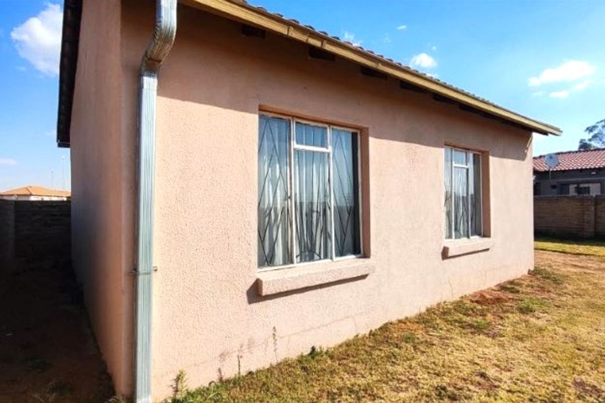 Rear view of the house featuring plastered walls, metal downpipe, and security-fitted windows, set within a fenced yard area.