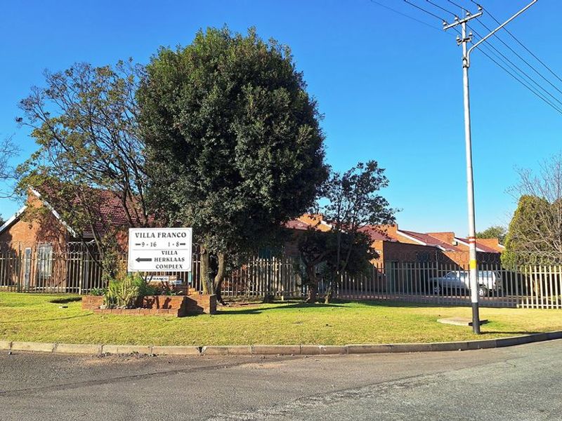 Street view of the Villa Franco apartment complex, featuring brick exterior walls, tiled roofing, and perimeter security fencing.