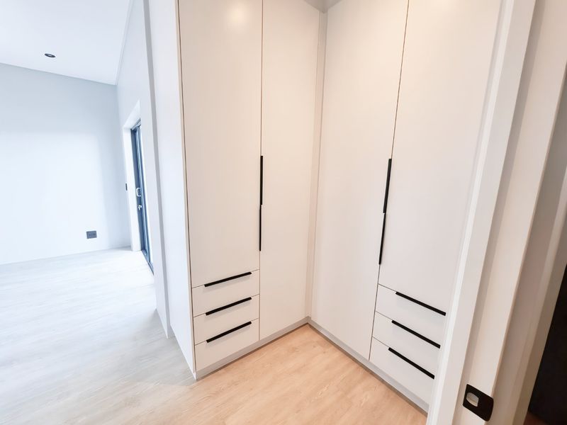 A bright, modern bedroom featuring sleek white built-in cupboards with black hardware, light wood-look laminate flooring, and access to an outdoor area.