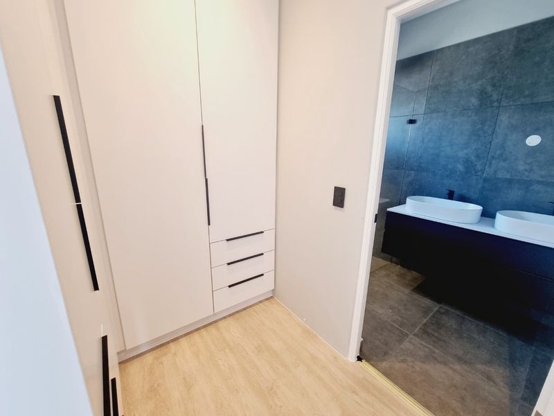 A modern hallway featuring built-in white cabinetry with sleek handles, leading into a contemporary bathroom with a double vanity and dark tiled finishes.