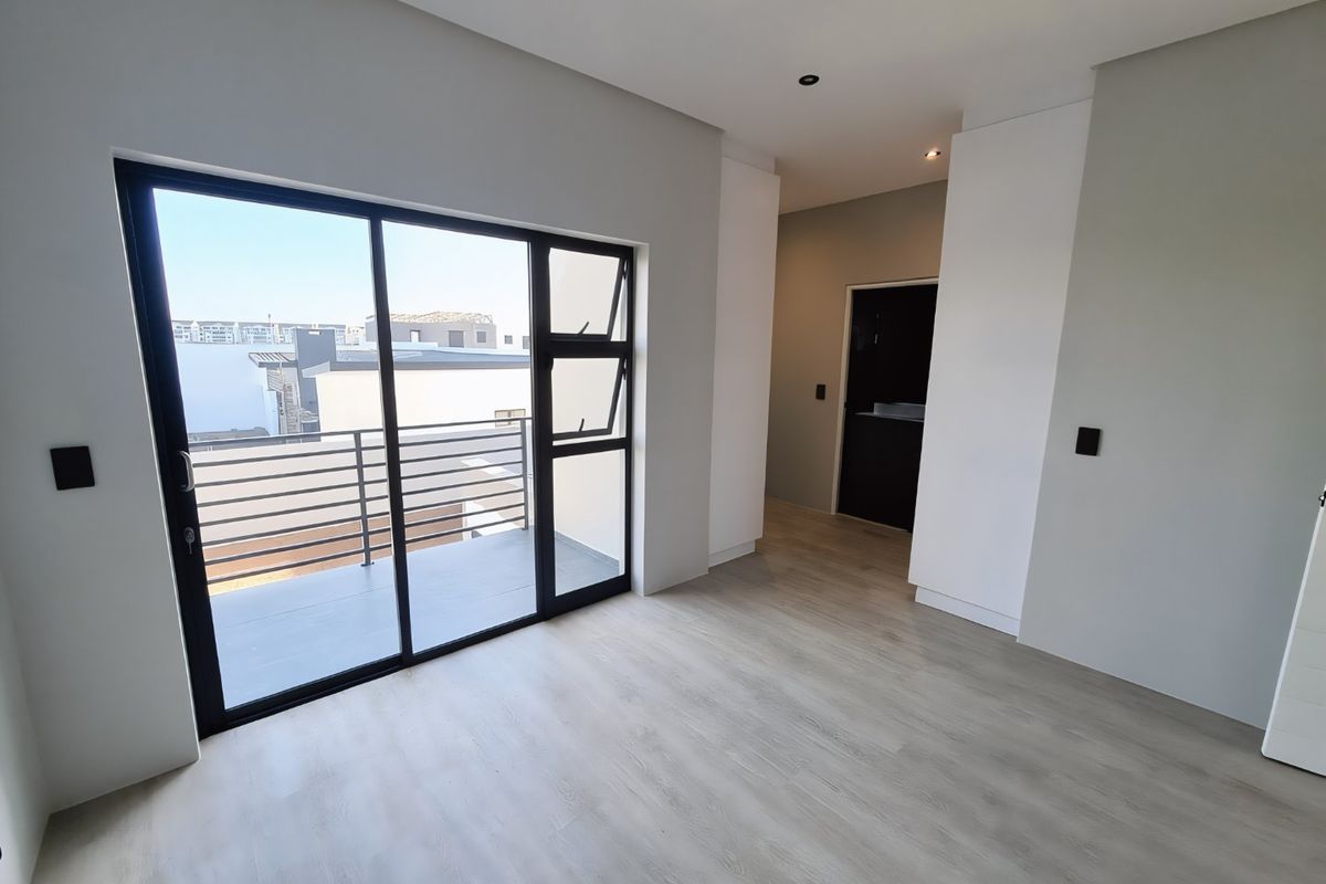 A bright, modern, empty bedroom featuring light laminate flooring, built-in cupboards, and a sliding glass door leading to a private balcony.