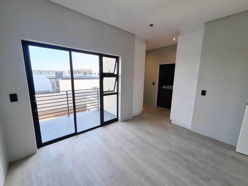 A bright, modern, empty bedroom featuring light laminate flooring, built-in cupboards, and a sliding glass door leading to a private balcony.