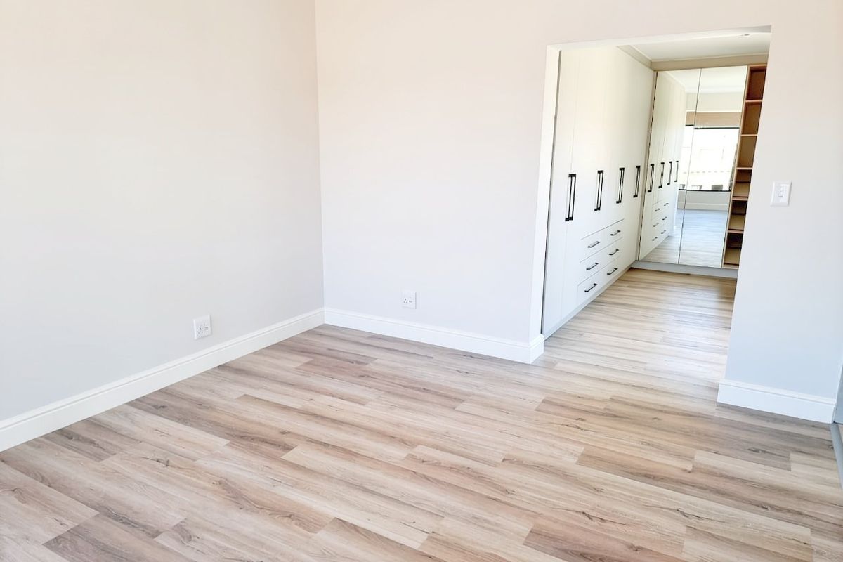 A bright, empty bedroom featuring modern light-toned laminate flooring and a spacious walk-in closet area with built-in cupboards and mirrored doors.