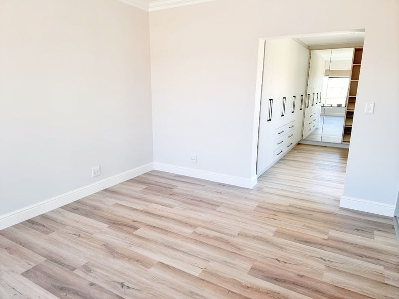 A bright, empty bedroom featuring modern light-toned laminate flooring and a spacious walk-in closet area with built-in cupboards and mirrored doors.