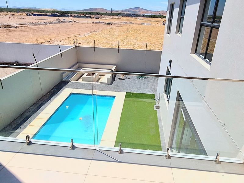 Rear exterior view featuring a swimming pool, artificial lawn, paved patio area with a built-in fire pit, and modern glass balustrade.