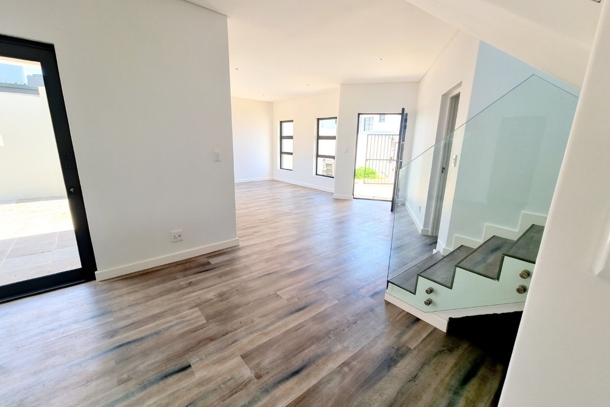 A bright, modern open-plan living area featuring wood-look laminate flooring, a contemporary glass balustrade staircase, and large aluminium-framed windows and doors.