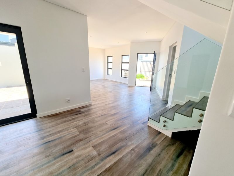 A bright, modern open-plan living area featuring wood-look laminate flooring, a contemporary glass balustrade staircase, and large aluminium-framed windows and doors.
