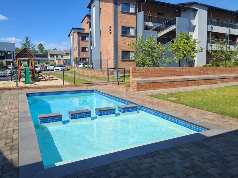 Communal swimming pool area featuring paved surrounds, brick boundary walls, and an adjacent playground within a well-maintained apartment complex.