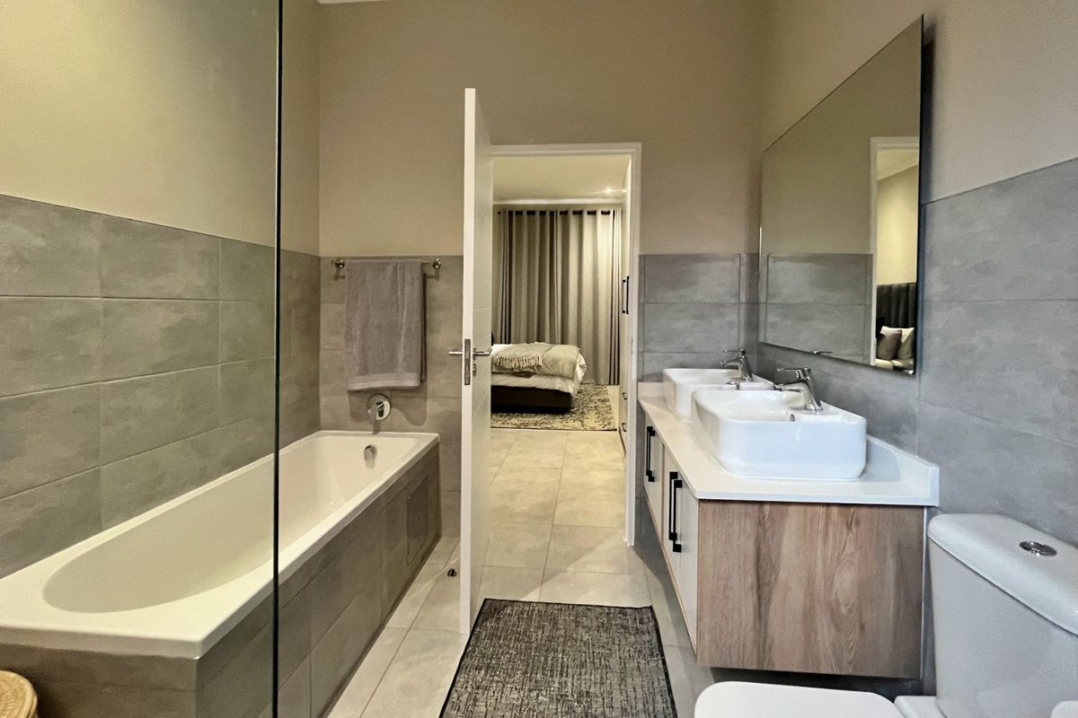 Modern bathroom featuring a double vanity with wood-finish cabinetry, a built-in bathtub with a glass screen, and neutral wall and floor tiling.