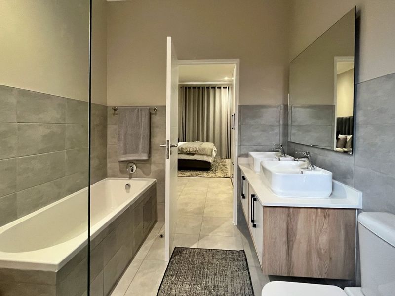 Modern bathroom featuring a double vanity with wood-finish cabinetry, a built-in bathtub with a glass screen, and neutral wall and floor tiling.