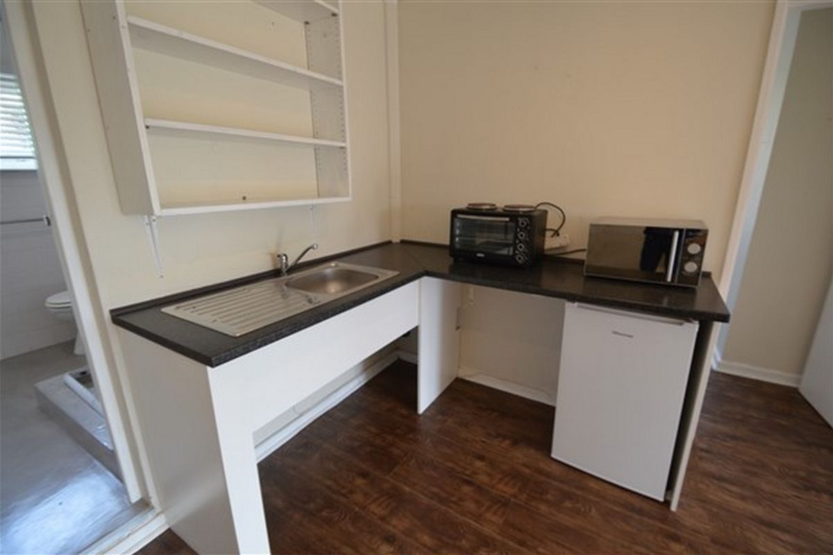 A compact kitchenette featuring a stainless steel sink, laminate countertop, and wall-mounted shelving, with direct access to an adjacent bathroom.