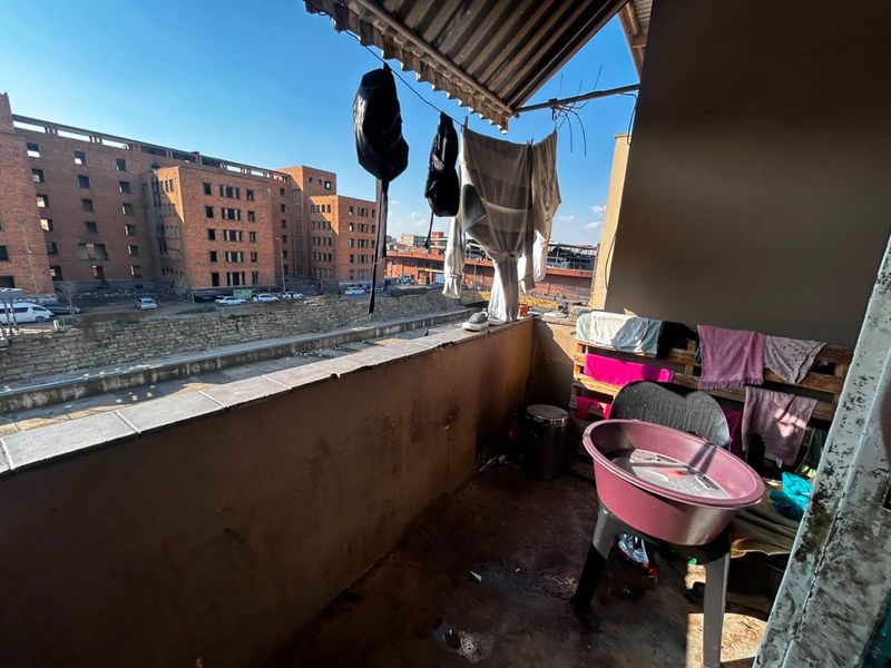 A small, weathered balcony featuring a corrugated metal roof and concrete balustrade, offering an urban view of the surrounding neighborhood.