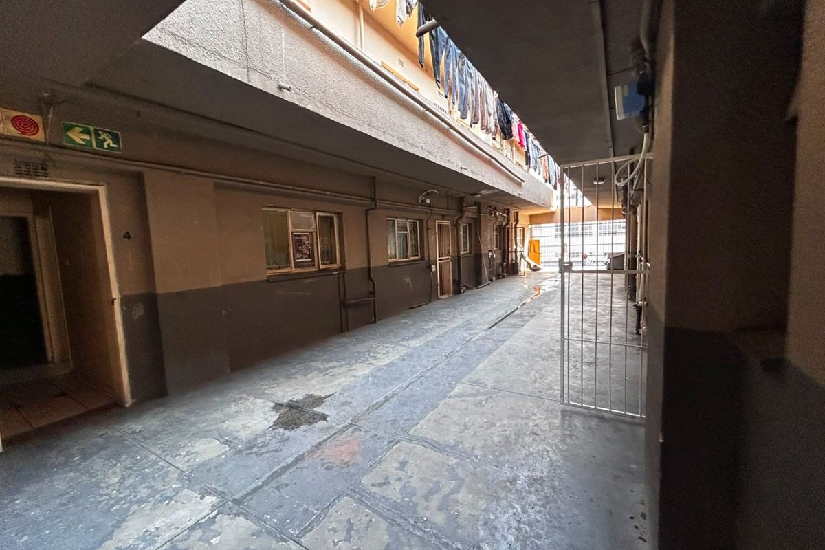 An exterior apartment building corridor featuring concrete flooring, security gates, and exposed utility piping along the walls.