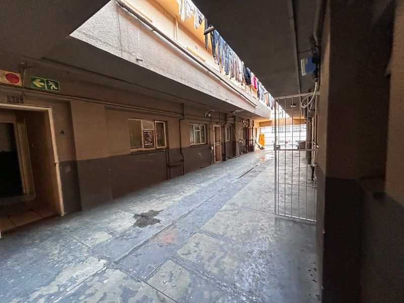 An exterior apartment building corridor featuring concrete flooring, security gates, and exposed utility piping along the walls.