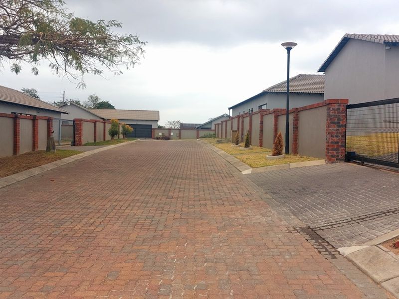 A view of a paved residential street within a complex, featuring brick boundary walls, secure sliding gates, and established street lighting.
