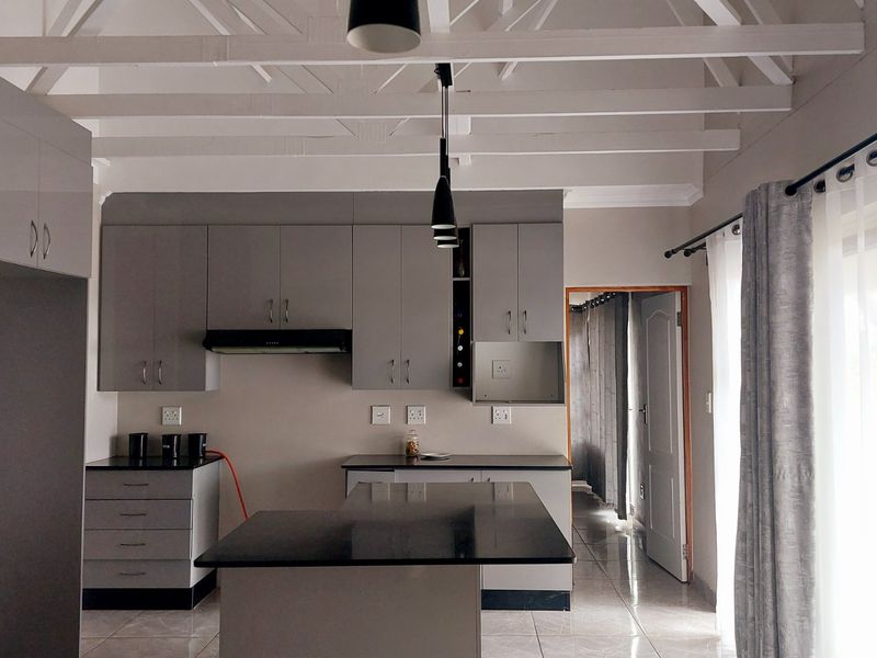 Modern kitchen featuring grey cabinetry, black granite countertops, exposed white timber ceiling beams, and tiled flooring with direct access to an adjacent hallway.