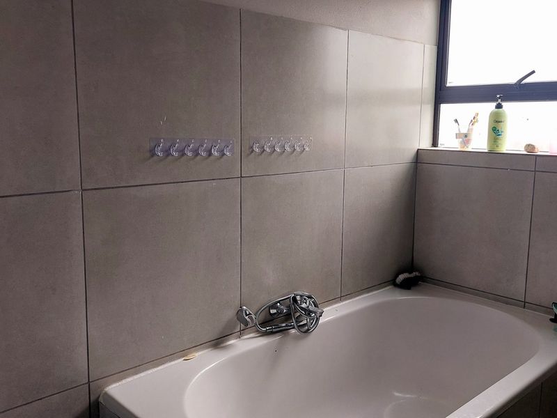 A functional bathroom featuring a built-in bathtub with chrome mixer tap and neutral grey wall tiling, complemented by a window for natural light.