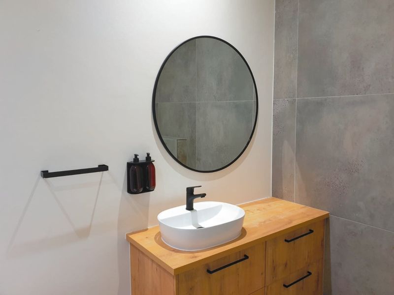 Modern bathroom vanity featuring a wooden cabinet, white vessel sink with black faucet, and a contemporary round mirror against large format grey wall tiles.