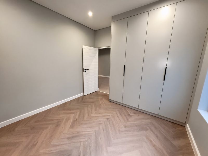A modern, empty bedroom featuring light grey walls, built-in cupboards, and high-quality herringbone vinyl flooring.