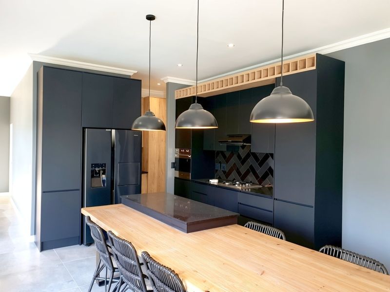 Modern kitchen featuring dark matte cabinetry, stone countertops, herringbone backsplash, integrated appliances, and a large wooden breakfast bar with pendant lighting.