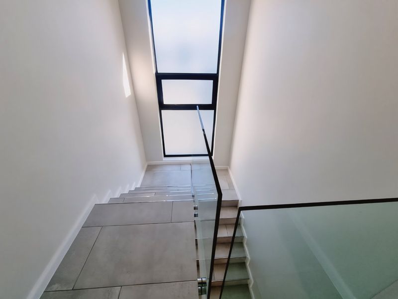 A modern staircase featuring large format floor tiles, a sleek glass balustrade, and a tall window providing ample natural light.