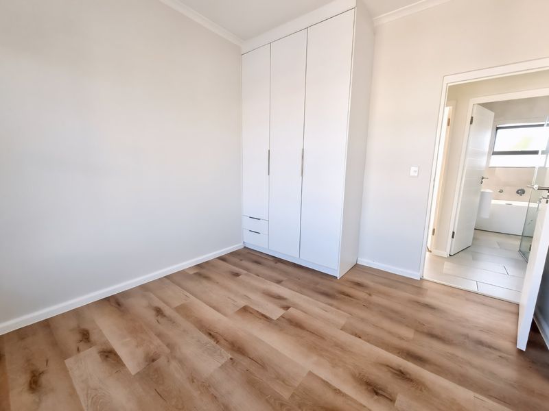 A bright, empty bedroom featuring modern laminate flooring and a built-in white wardrobe, with an adjacent en-suite bathroom visible through the doorway.