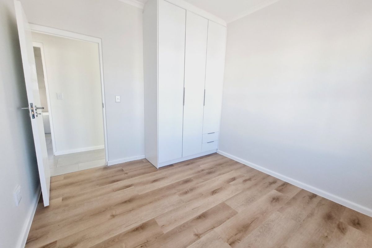 A bright, empty bedroom featuring modern laminate flooring, neutral walls, and a clean, white built-in wardrobe with integrated drawers.