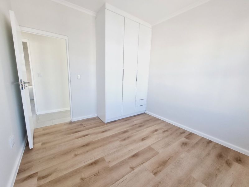 A bright, empty bedroom featuring modern laminate flooring, neutral walls, and a clean, white built-in wardrobe with integrated drawers.