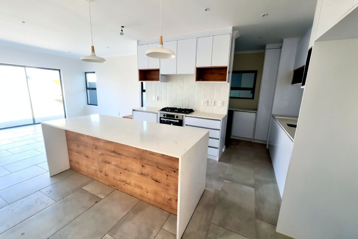 Modern open-plan kitchen featuring a large island with wood-grain detailing, quartz countertops, gas hob, and herringbone backsplash, flowing into a bright living area.