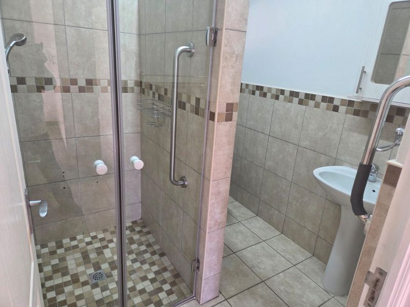 A functional bathroom featuring a glass-enclosed shower, pedestal basin, and tiled finishes with installed safety grab rails.
