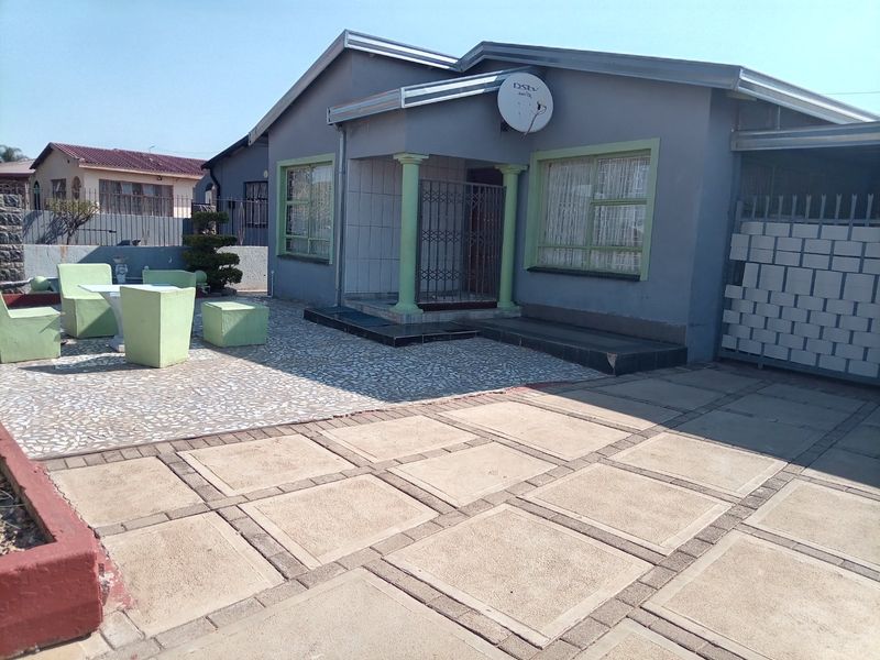 Front exterior view of a single-story house featuring a paved driveway, tiled patio area, and secure entrance with metal gate.