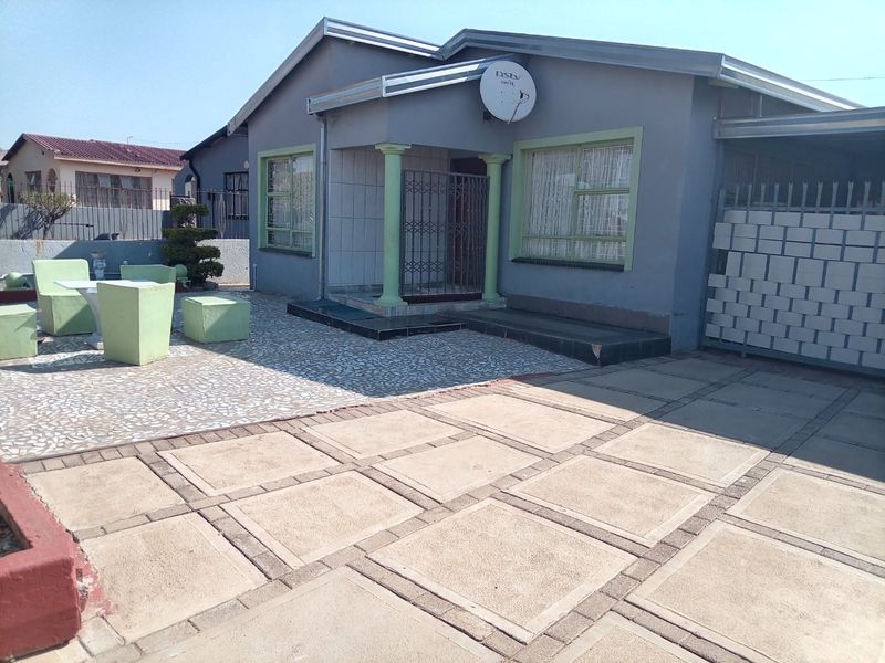 Front exterior view of a single-story house featuring a paved driveway, tiled patio area, and secure entrance with a metal security gate.