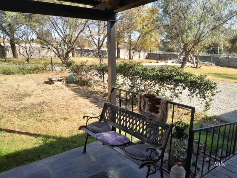 A covered patio area featuring slate tile flooring and a metal railing, overlooking a spacious garden with mature trees.