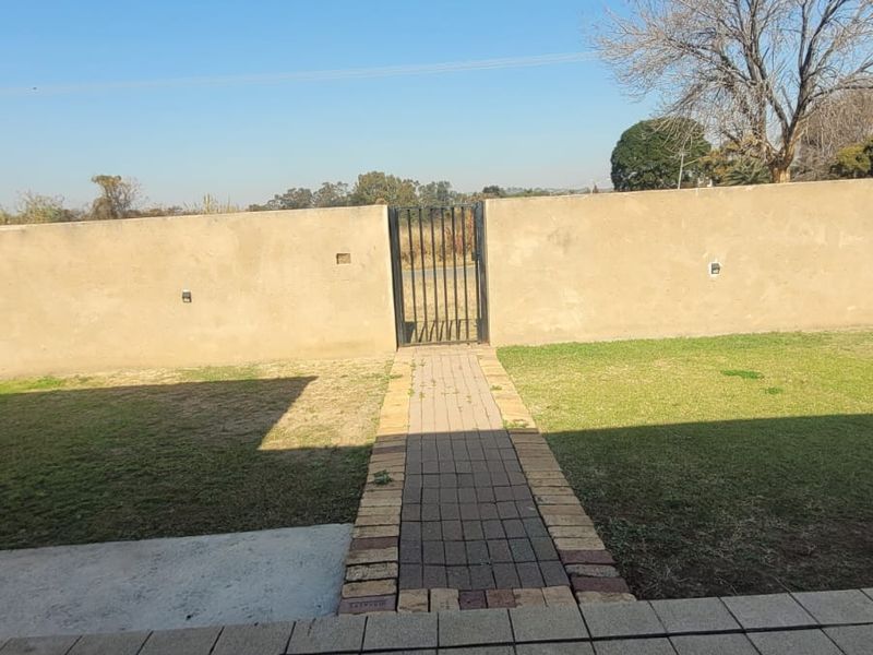 A rear outdoor area featuring a paved patio, a brick pathway leading to a metal gate, and a perimeter wall enclosing the lawn.