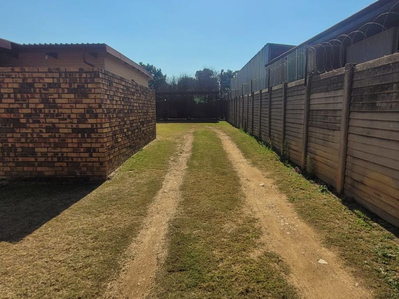 Rear exterior view showing a dirt driveway between a brick structure and a precast concrete boundary wall topped with razor wire.